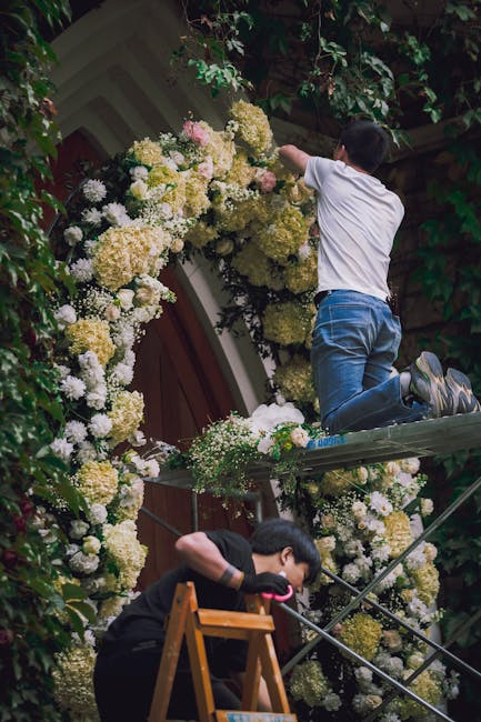 Two children are engaged in decorating a floral archway outside a building with lush green foliage. One child, dressed in a white t-shirt and blue jeans, is kneeling on a small scaffold, reaching up t