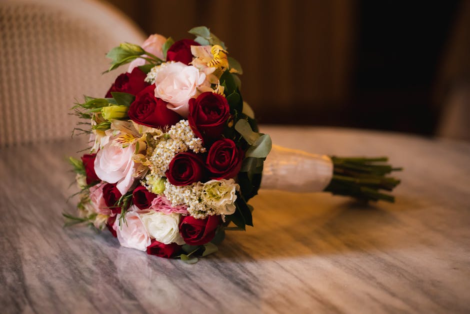 A bouquet arrangement of fresh flowers resting on a wooden surface, featuring a mix of deep red roses, soft pink and white ranunculus, delicate white and yellow alstroemeria, and clusters of small whi