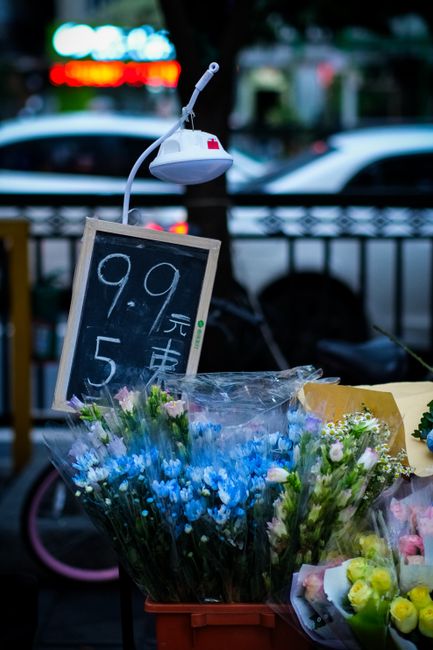 A vibrant display of fresh flower bouquets arranged outdoors on a market stall, featuring a mix of blue, pink, and white flowers with green foliage. The flowers are wrapped in clear plastic and set in