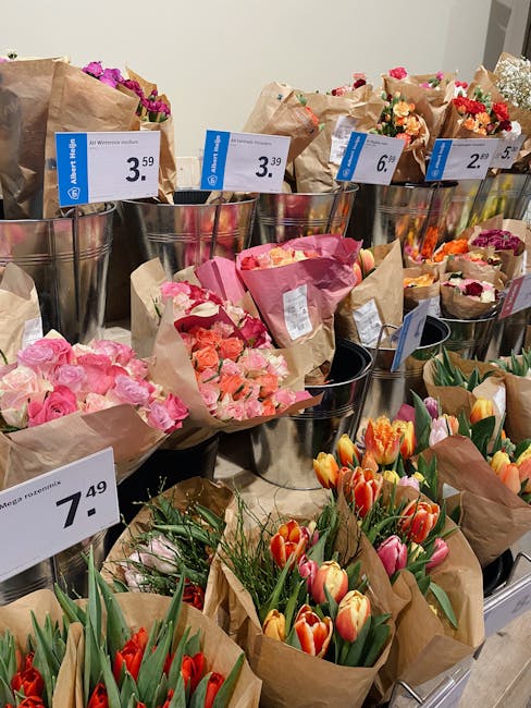 A display of various flower bouquets arranged in metallic buckets and wrapped in brown craft paper at a flower shop. The arrangements include pink roses, peach and pink carnations, orange and pink tul