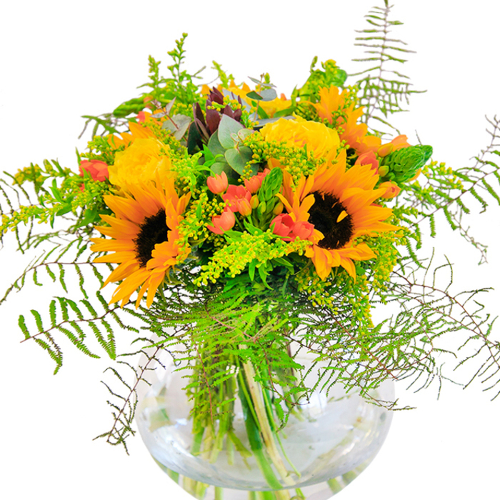 The initial impression of this Archway-crafted bouquet is a rush of light and joy-a hand-tied symphony where golden sunflowers and lush yellow garden roses stand side by side. Sunflowers, proud and sunlit, radiate from the centre, each petal perfectly unfurled to frame the classic, swirling roses whose buttery layers seem to glow from within. Rich green leaves and trailing vines sweep outward, creating a halo of freshness akin to walking under the leafy canopies of Dartmouth Park. Tucked between, clusters of scarlet hypericum berries sparkle with life, their gleaming surfaces contrasted with soft, matte petals. A variety of textured greenery stretches outward, from airy ferns to fine, flower-tipped sprigs of goldenrod, all balanced by subtle eucalyptus leaves that introduce notes of calming blue-green. The round glass vase reveals every stem and root, grounding the arrangement in honest, organic beauty, while reflected light brightens each bloom and leaf. Up close, you sense the interplay of velvet petals, crisp greenery, and the bouquet's airy scent-reminding one of summer gardens and cherished gatherings along Archway's historic streets. Whether for a special occasion or a spontaneous gesture, this bouquet marks the moment with exuberant colour and gentle nostalgia.