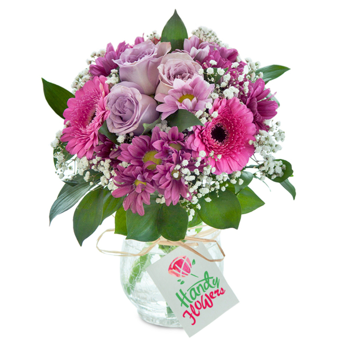 A stunning bouquet, artfully arranged in Archway and ready for thoughtful delivery, greets you from its clear glass vase with a subtle crackled texture reminiscent of city windows kissed by rain. At the core, tender lavender roses unfurl with elegance-each petal a brushstroke of delicate romance and gentle fragrance. Their beauty is amplified by vivacious fuchsia gerbera daisies, their broad petals and dark central eyes catching every ray of light much like the bustling vibrancy of Archway Market. Drawing the eye further are clusters of deeply coloured magenta chrysanthemums, with some yellow-green centers sparkling lively interest into the arrangement. Sprigs of baby's breath-tiny, white, and cloud-like-bring airy contrast and a sense of playful lightness to the ensemble. The bouquet's lush, deep green leaves wrap the blossoms in effortless grace, echoing the greenery of Waterlow Park nearby. A rustic touch is added by a length of light brown raffia, tied in a bow around the vase's neck, from which a classic white gift tag hangs, prepared for heartfelt greetings. This radiant mix is a tribute to the rich character of Archway, bringing together colour, freshness, and professional craftsmanship for any memorable occasion or everyday joy.