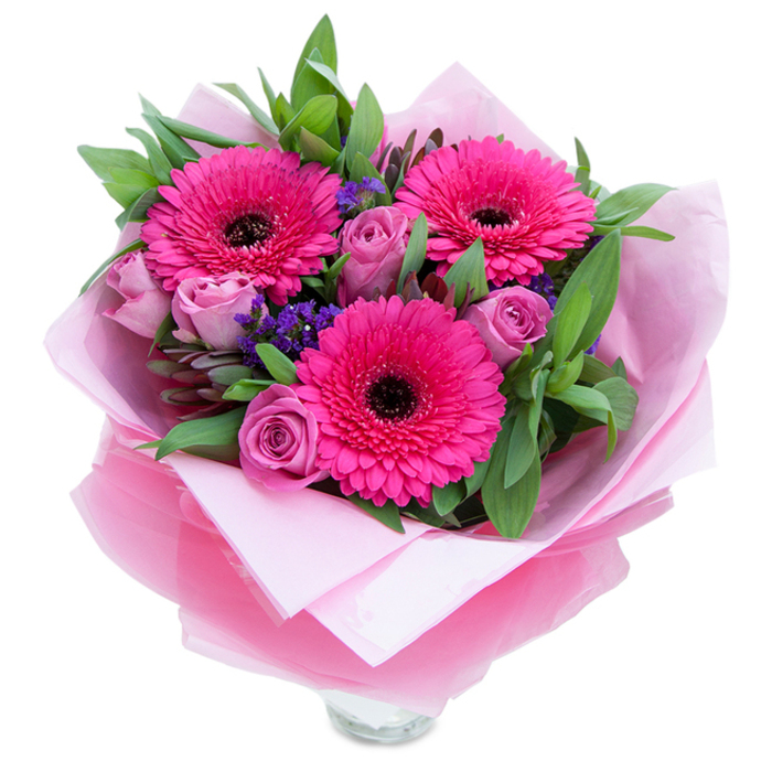 There is a playful, graceful energy to this bouquet: three fuchsia Gerbera daisies open wide like small, jubilant suns, their deep centres providing a striking contrast to the vibrant petals. Nestled among them, roses in a softened lavender-pink or dusty-rose tone curl inward in velvety layers, their texture inviting the fingertips to linger. Sprays of petite purple statice scatter between the roses and gerberas, introducing a fine, twinkling texture that resembles wildflowers tucked into a cultivated posy. The piece is robed in layers of packaging that feel both luxurious and intimate - an inner matte pale-pink tissue gives a muted, satin cushion to the stems, while an outer translucent fuchsia-pink cellophane, slightly crinkled, adds a shimmering veil and reinforces the bouquet's pink identity. Wide green leaves, reminiscent of salal or eucalyptus, surround the blooms, adding structure, cool colour contrast, and a hint of crisp, dewy foliage scent. The top rim of a clear glass vase is visible at the base, suggesting the bouquet will sit elegantly on a mantel or dining table in an Archway home or a flat near Tufnell Park. In early daylight the bouquet feels fresh and optimistic; the roses breathe a gentle perfume, the gerberas a mild vegetal sweetness, and the statice gives a subtle, dried-floral note. This arrangement is designed to celebrate - birthdays, anniversaries, a new home on Junction Road - or simply to lift a neighbour's day with bright, considerate beauty.