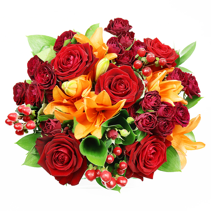 Captured from above on a plain white surface, this densely packed, circular bouquet presents an arresting study in warm tones and varied textures. Large, deep-red roses dominate the scene, their velvety petals unfurling in rich, saturated layers that evoke classic romance and an intimate warmth. Interspersed among them are radiant orange Asiatic lilies - some explosively open with their stamens prominent, others in the graceful arc of buds about to bloom - introducing bright trumpet shapes that energise the composition. Smaller spray roses in a wine or burgundy shade nestle into the spaces between the major flowers, their compact clusters adding visual depth and a luxurious, tactile feel. Glossy hypericum berries in vivid ruby-red form gleaming, spherical accents that punctuate the bouquet and play with the light. Cool, broad green leaves, likely ruscus or salal, peek and fan out around the flowers, providing structural support and a fresh contrast that lifts the warm palette. The lighting is even and gentle, chosen to show colour saturation and texture rather than dramatic shadow, so each petal's surface - from the soft nap of the roses to the silky sheen of the lilies - is clearly legible. The overall effect is sumptuous and joyful: a bouquet that speaks of heartfelt celebration, from anniversaries to milestone birthdays, and that would travel beautifully for same-day delivery across Archway and nearby Holloway Road addresses. As a local florist I imagine it brightening a mantel in a Dartmouth Park home or arriving as a vivid, romantic gesture along Junction Road.